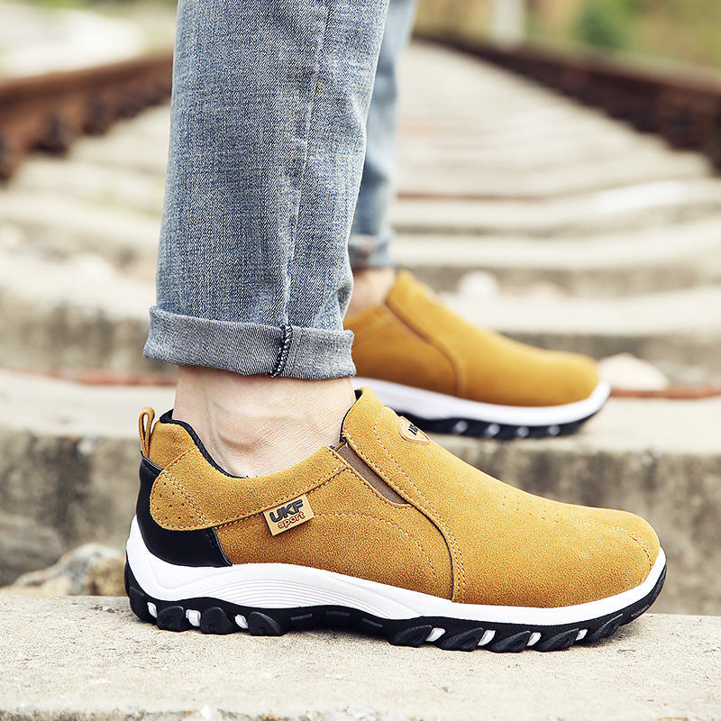Man wearing yellow suede mountain shoes outdoor men's shoes lazy shoes with black and white soles, paired with rolled-up denim jeans on a stone surface near train tracks.